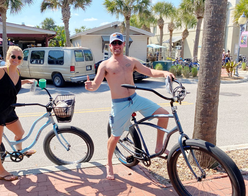 Two cyclists smiling while riding hybrid bikes on a beachside path.